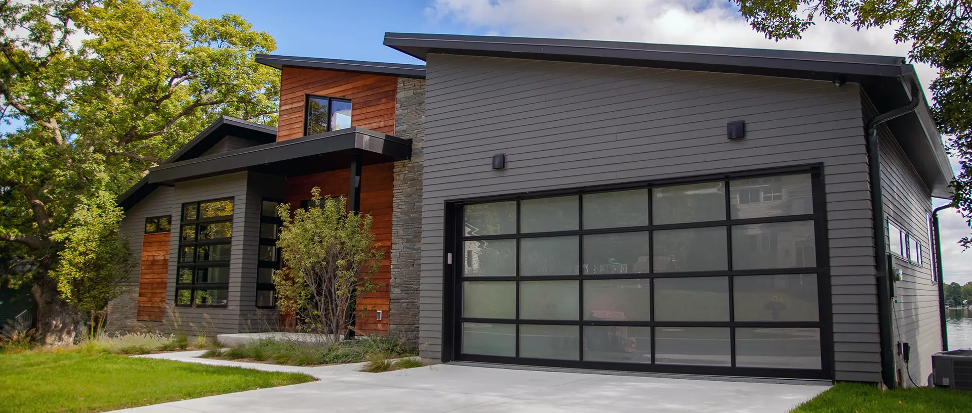 Modern home featuring a black aluminum-frame garage doors with glass panels, in front of a lake.