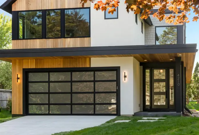 Modern house with wood and white exterior. Features large glass windows, a black framed garage, and a sleek front door. Lush greenery surrounds.